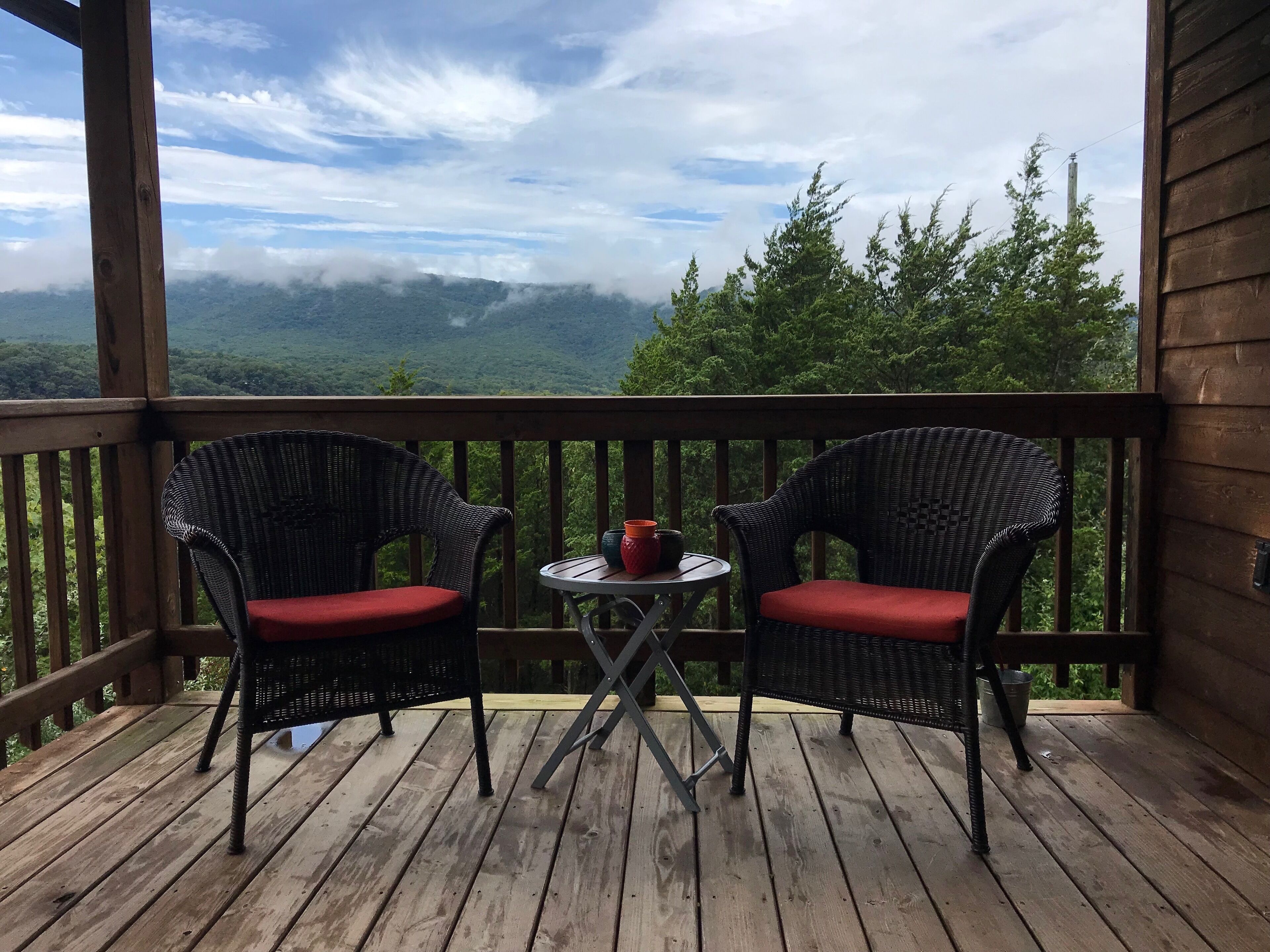 A wooden deck with two wicker chairs and a small table, overlooking a forested mountain area.