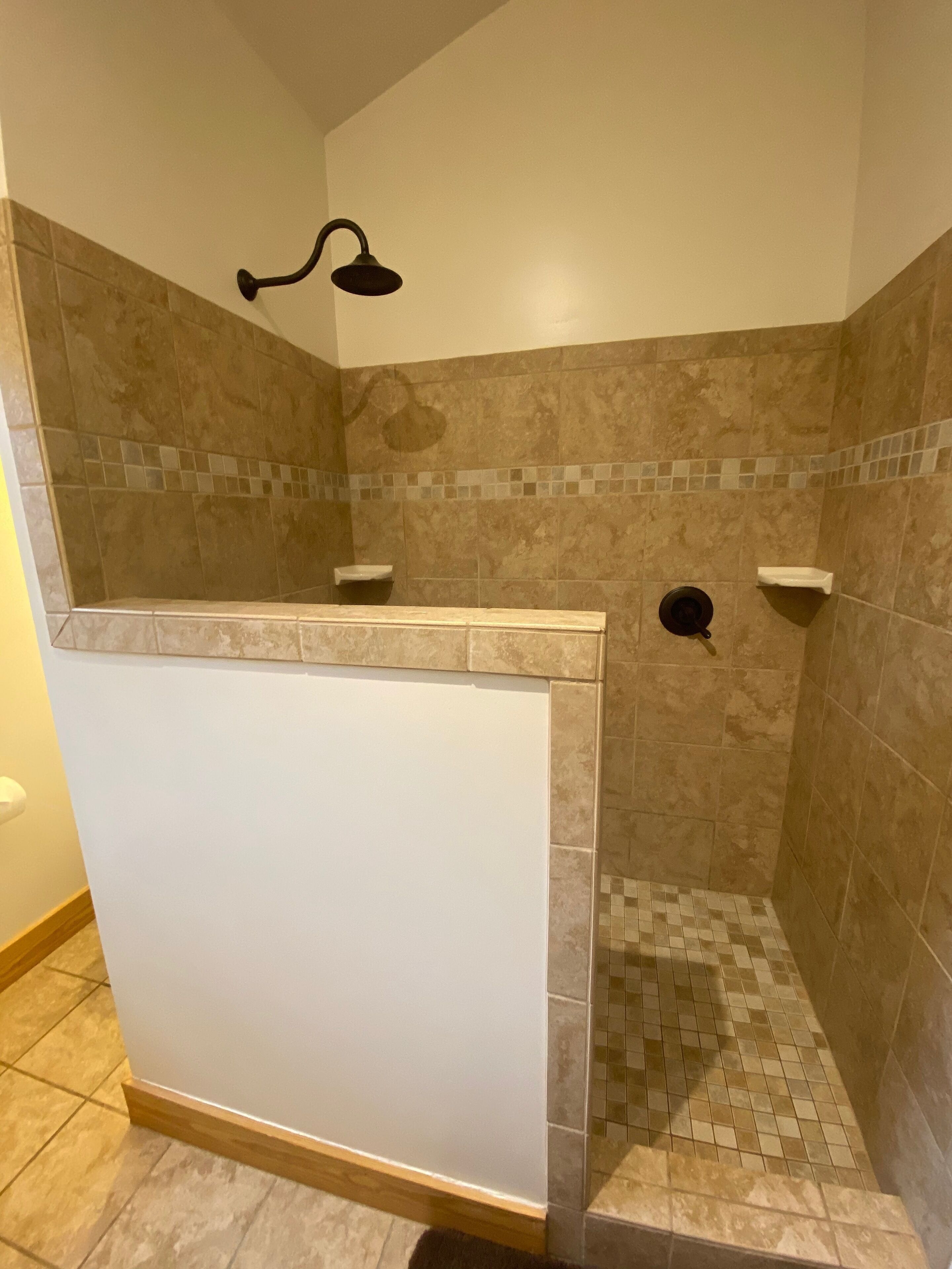 A shower with beige tiles, a white partition, and a handheld showerhead.