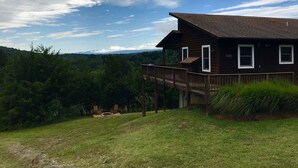 Una cabaña de madera con terraza, rodeada de vegetación exuberante y montañas al fondo.
