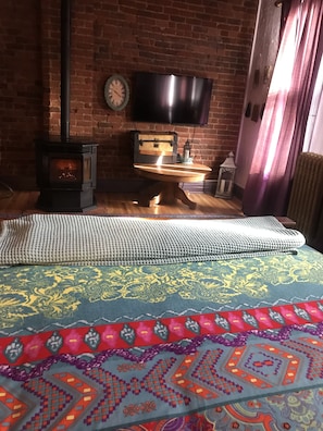Una habitación acogedora con pared de ladrillo, suelo de madera, cama con ropa de cama estampada, una mesita con un farol, y un televisor montado en la pared.