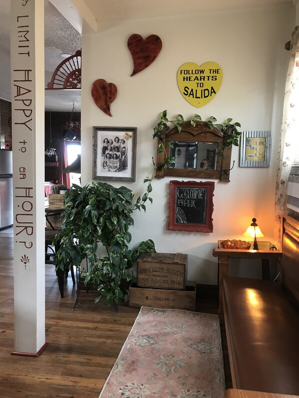 Una habitación acogedora con suelo de madera, una planta verde grande, una pizarra con la inscripción 'BIENVENUE FRANK' y un cartel que dice 'SIGUE LOS CORAZONES A SALIDA'.