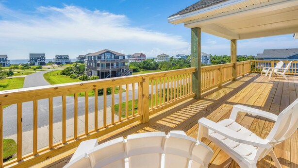 A wooden deck with white plastic chairs, overlooking a coastal residential area with houses and a road. - Soundside Dog Friendly Home w/ Pool, Hot Tub, Game Room, Community Boat Ramp (Salvo)