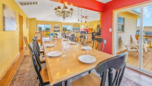 A dining area with a long wooden table set for six, surrounded by chairs, with a view of a balcony and a kitchen in the background. - Soundside Dog Friendly Home w/ Pool, Hot Tub, Game Room, Community Boat Ramp (Salvo)