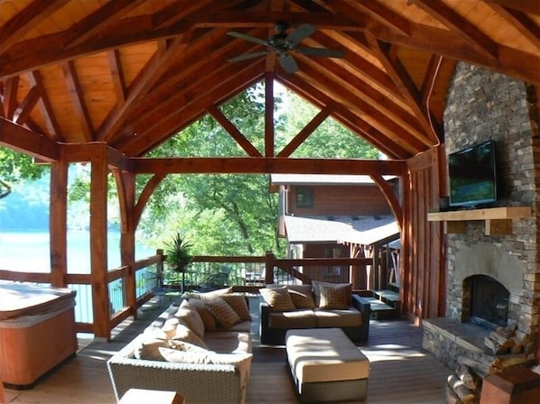A covered outdoor seating area with wooden furniture, a stone fireplace, and a television mounted on the wall.