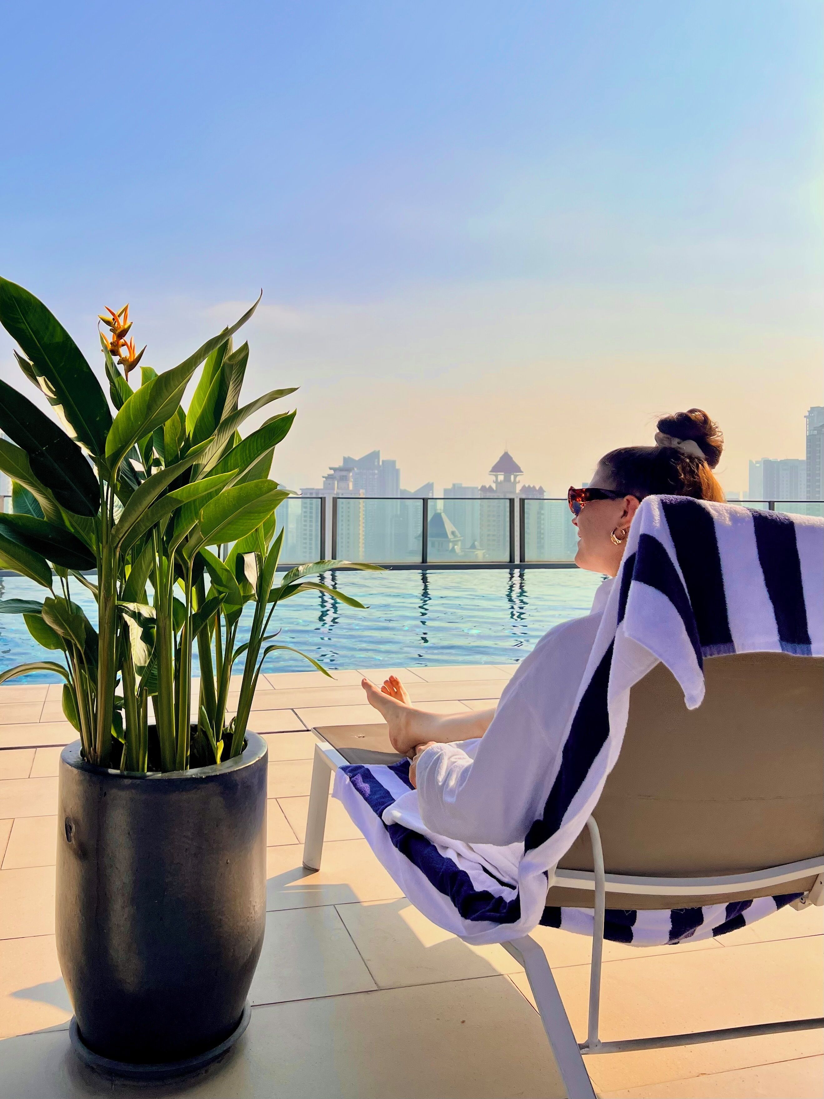 Eine Person entspannt in einer Liege am Pool, im Hintergrund ist eine Stadtlandschaft zu sehen.