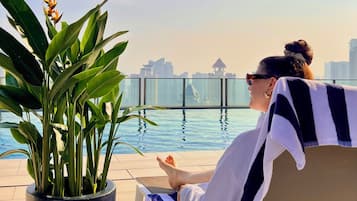 Una persona descansando en una silla de salón junto a una piscina, con un horizonte urbano de fondo.