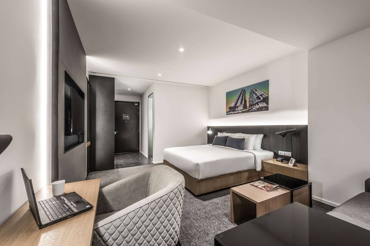 Una habitación de hotel moderna con una cama, un escritorio con una computadora portátil, una silla, una mesita de noche y un cuadro en la pared.