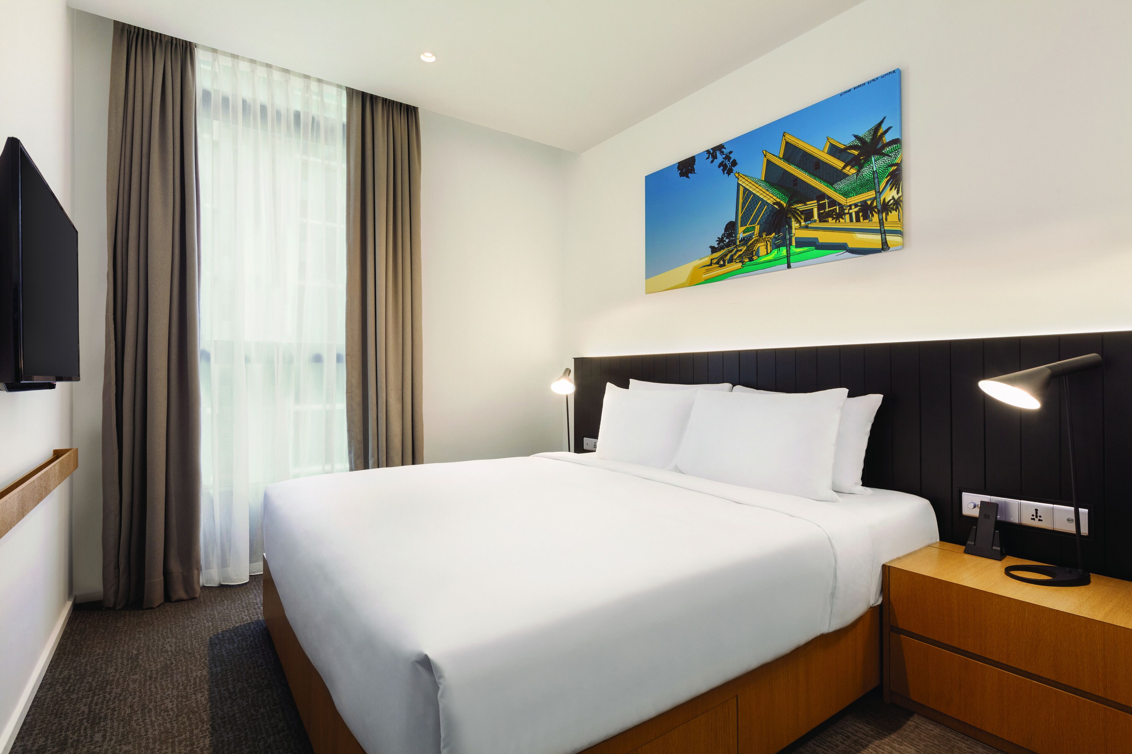 A hotel room with a large bed, a headboard, a bedside table with a lamp, a wall-mounted TV, and a painting of a building.