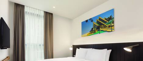 Quarto de hotel com uma cama grande, cabeceira, mesa de cabeceira com abajur, televisão na parede e uma pintura de um edifício.