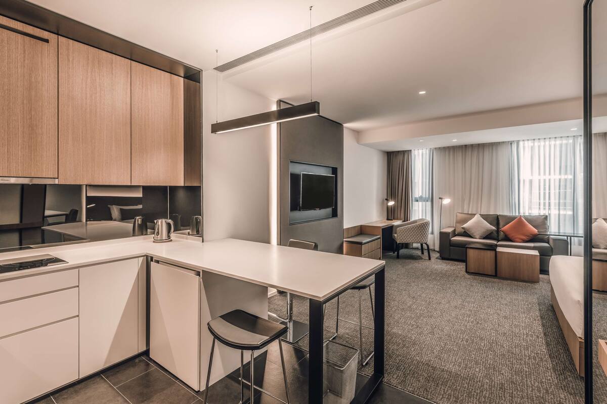 Una habitación de hotel moderna con kitchenette, zona de comedor y sala de estar con sofá y televisión.
