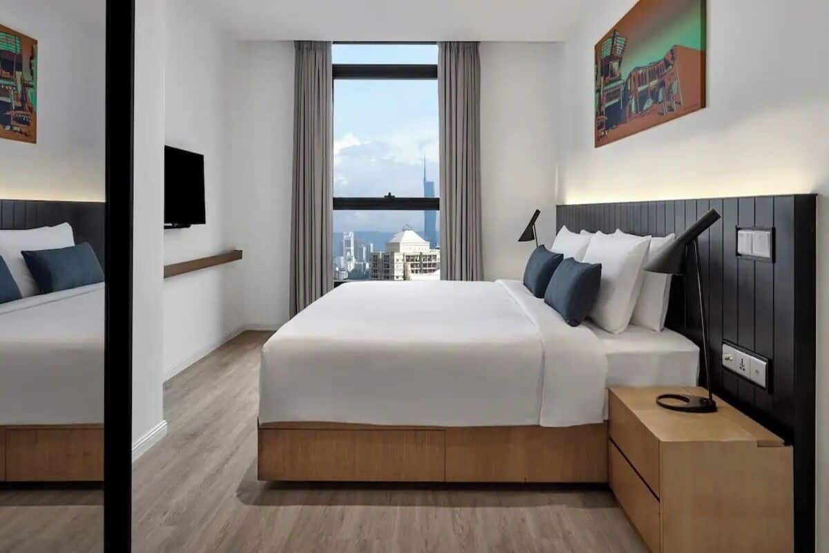 Una habitación de hotel moderna con una cama grande, una mesita de noche, un televisor y vista a la ciudad por la ventana.