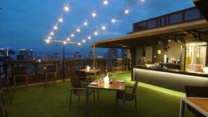 An outdoor dining area with tables and chairs, string lights, and a city view at dusk.