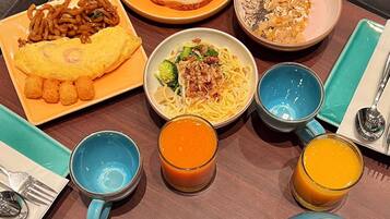 A table set with various dishes including noodles, an omelet, and fruit, along with two glasses of orange juice and a blue teapot.