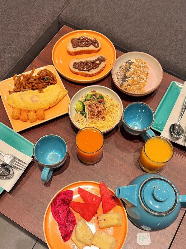 Una mesa servida con diversos platos, incluyendo fideos, una tortilla y fruta, junto con dos vasos de jugo de naranja y una tetera azul.