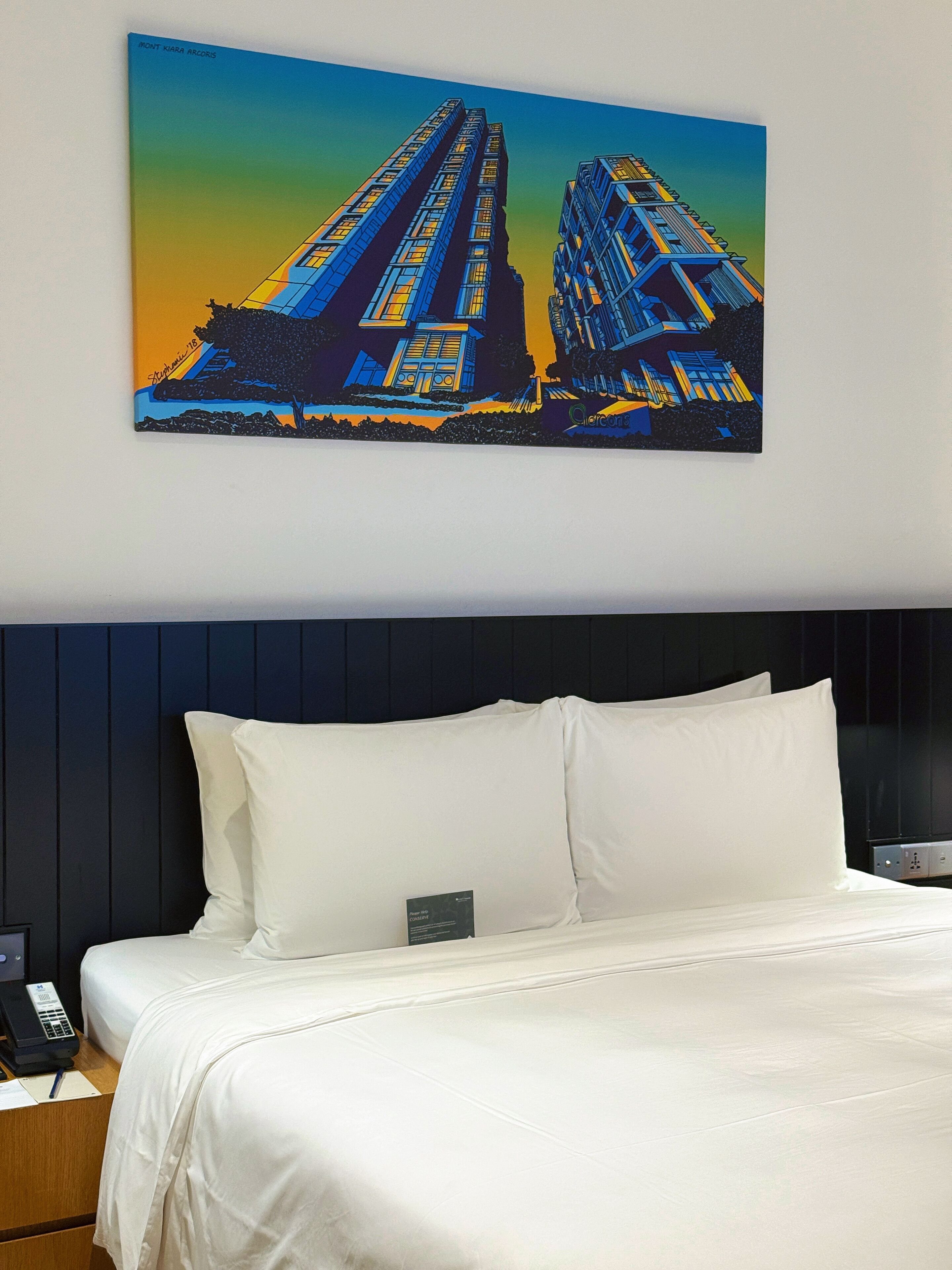 A hotel room with a large bed, a headboard, and a painting of buildings on the wall.