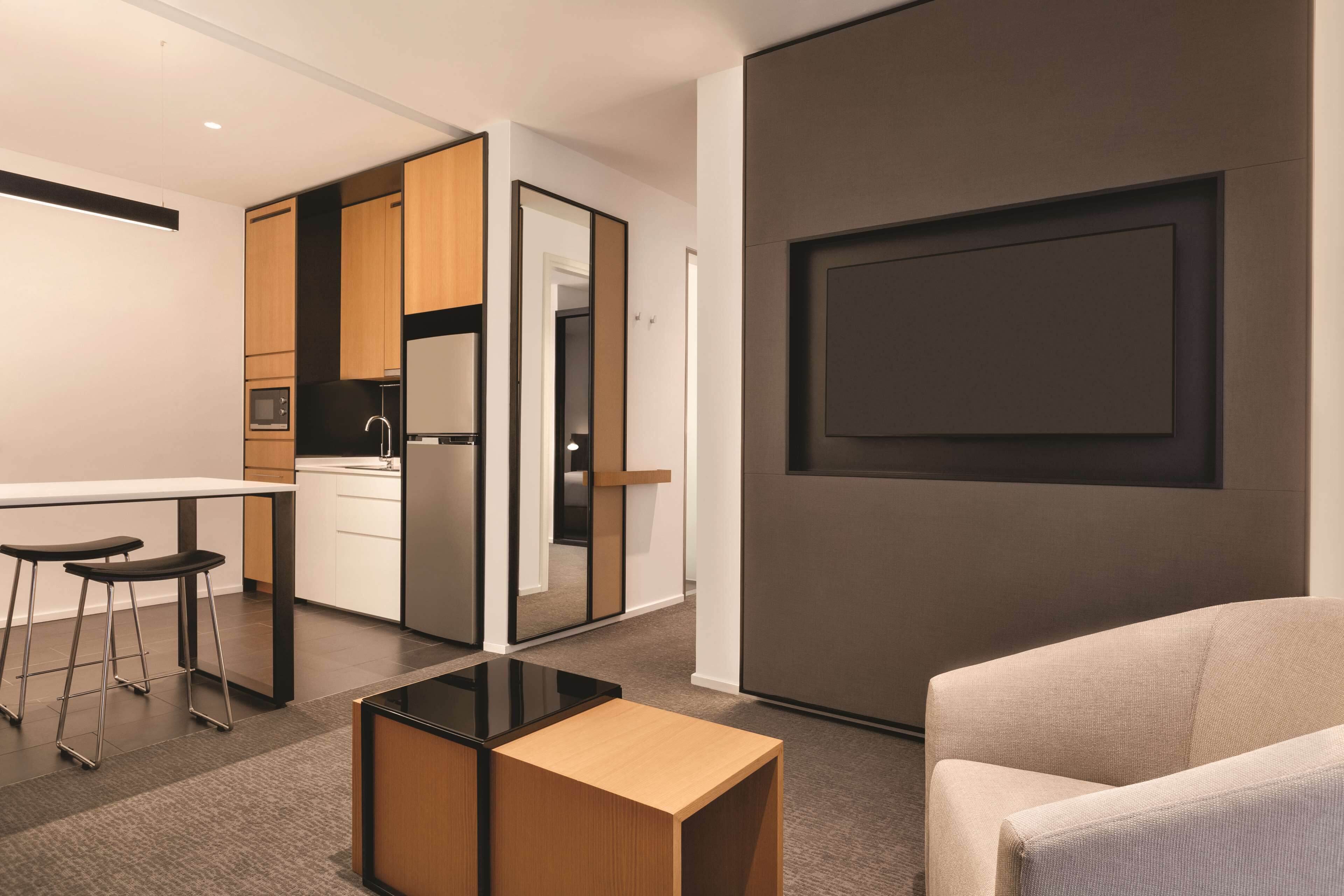 Een moderne keuken met een koelkast, magnetron en houten kastjes. Er is een eethoek met een tafel en twee barkrukken. Een grote spiegel is aan de muur bevestigd, en een bank is in de voorgrond zichtbaar.