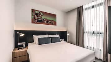 A hotel room with a large bed, two bedside tables, a nightstand, a painting above the bed, and a view through sheer curtains.