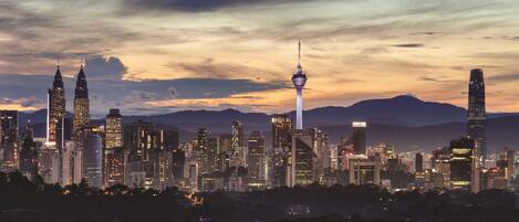 A city skyline at dusk with prominent skyscrapers, including the Petronas Towers and a modern tower.