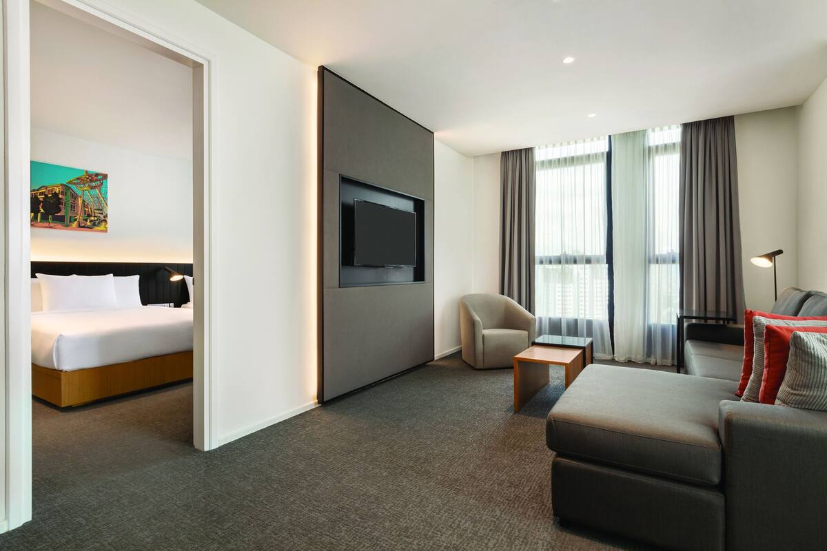 Una habitación de hotel moderna con una cama, un televisor montado en la pared, un área de descanso con un sofá y sillones, y una mesita.
