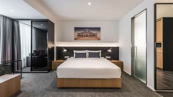 Et moderne hotelværelse med en stor seng, et billede over sengen, et skrivebord og en glasvæg, der adskiller værelset fra et spiseområde.
