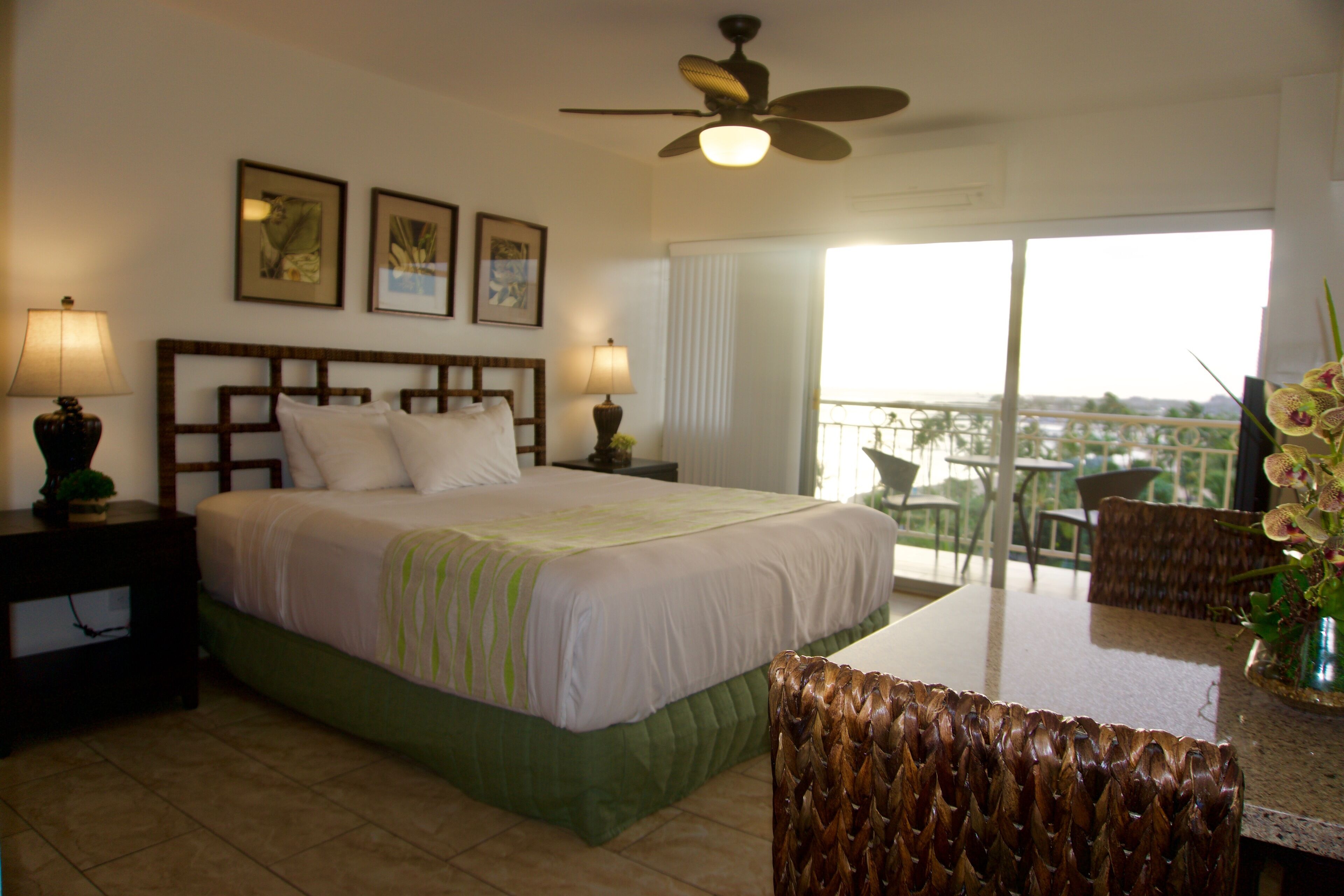 Quarto de hotel com cama, duas mesas de cabeceira com luminárias, ventoinha no teto, varanda com vista e área de jantar com cadeira e mesa.