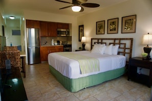 Quarto de hotel com cama, escrivaninha, cadeira, cozinha compacta e ventoinha no teto.