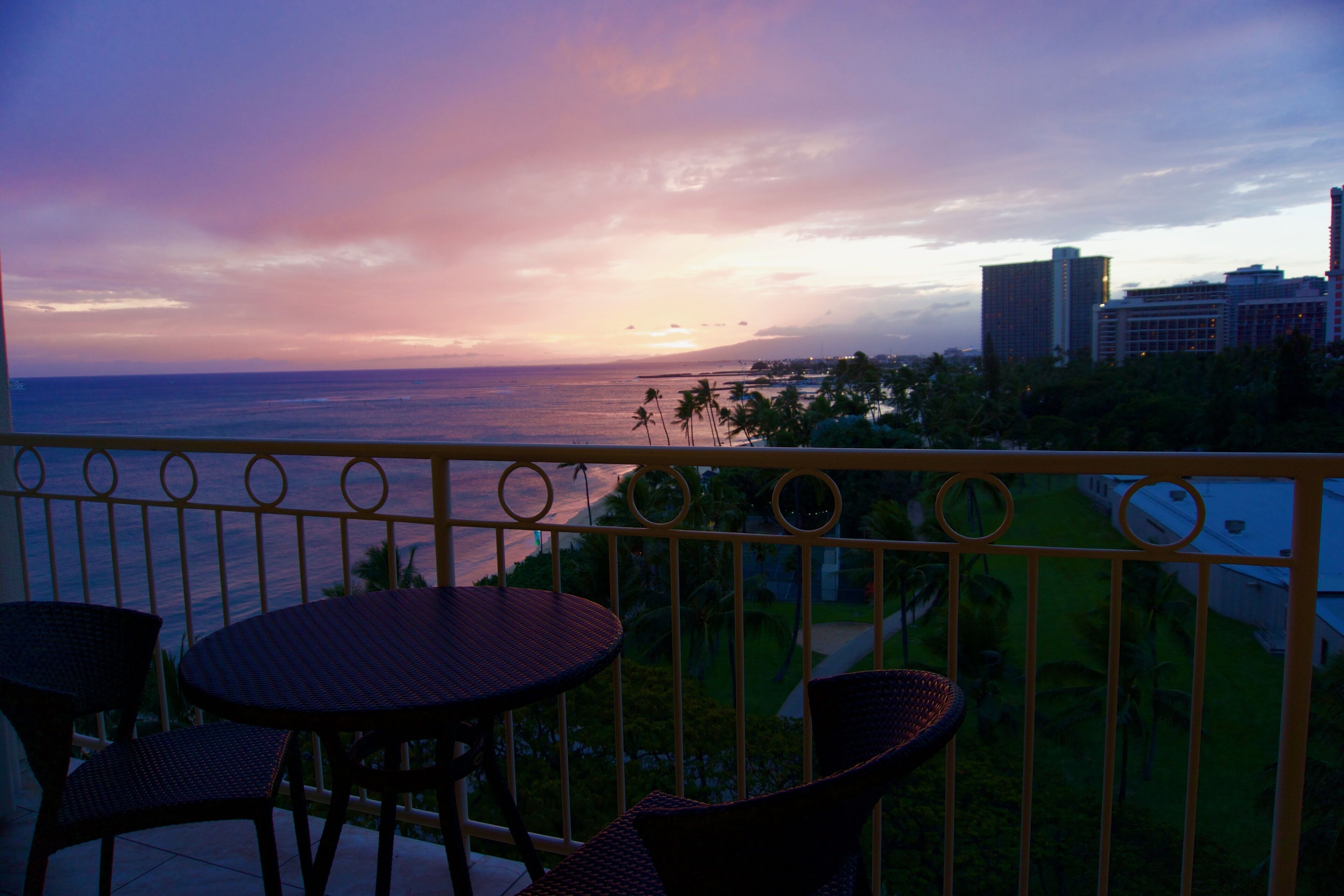 A balcony with wicker chairs and a table, overlooking a cityscape and ocean at sunset.