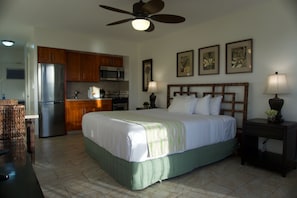 A hotel room with a bed, a nightstand, a lamp, a ceiling fan, a kitchen area, and a microwave. - Waikiki Beach ocean view studio with pool access right on the white sand beach. (Honolulu)