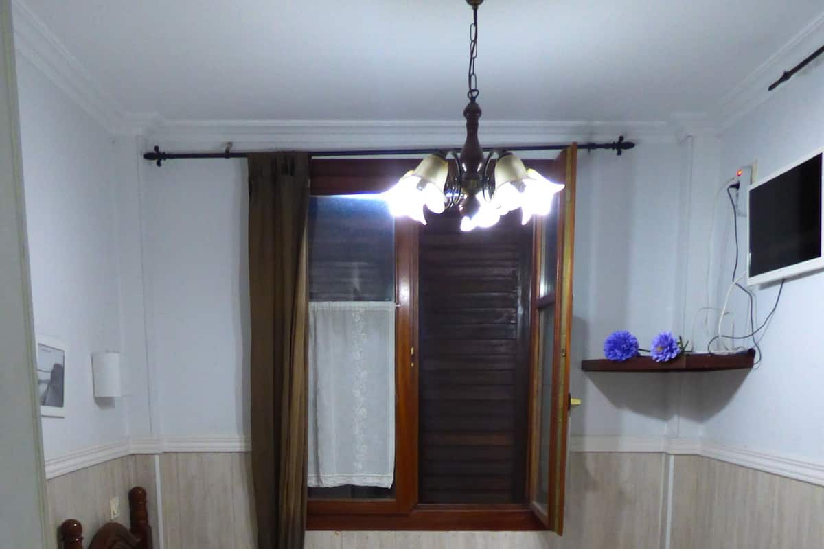 A room with two single beds, a wooden headboard, a small bedside table, a nightstand, a wall-mounted TV, a window with curtains, and a ceiling light fixture.