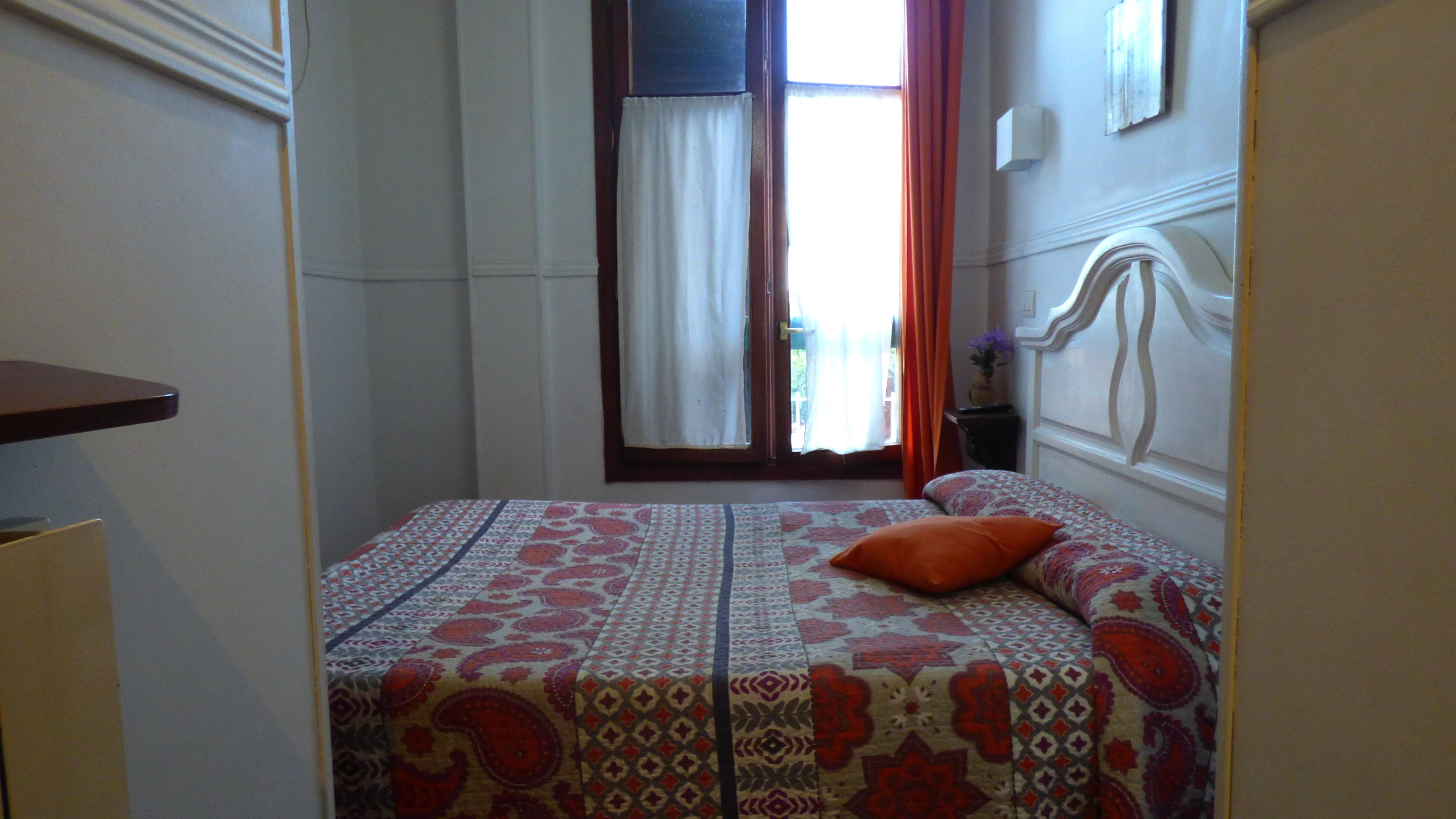 A bedroom with a bed, a nightstand, a window with curtains, and a view of the outdoors.