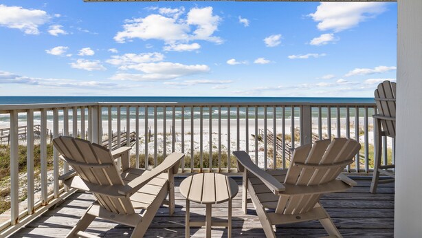 A wooden deck with two Adirondack chairs and a small table, overlooking a beach with a clear sky.