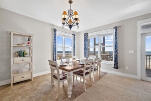 A dining room with a wooden table and white chairs, a chandelier, and a view of the ocean through large windows. - Happy Hours- 5 BR Home on N. Cape- Beachfront, Pet Friendly, Sleeps 16 (Cape San Blas)
