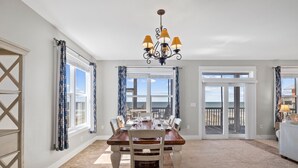 A dining area with a wooden table, chairs, and a chandelier. There are large windows with curtains, and a view of the ocean.