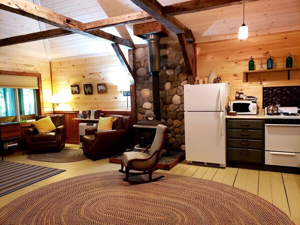 A cozy cabin interior with a stone fireplace, wooden beams, a kitchen area, and a seating area with a fireplace. - Cottage on the Kennebec (Caratunk)