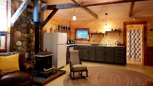 A rustic cabin interior with a stone fireplace, wooden beams, a kitchen with a large window, and a cozy seating area. - Cottage on the Kennebec (Caratunk)