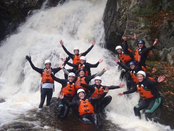 Un grupo de personas con chalecos salvavidas y cascos, de pie y sentadas frente a una cascada.