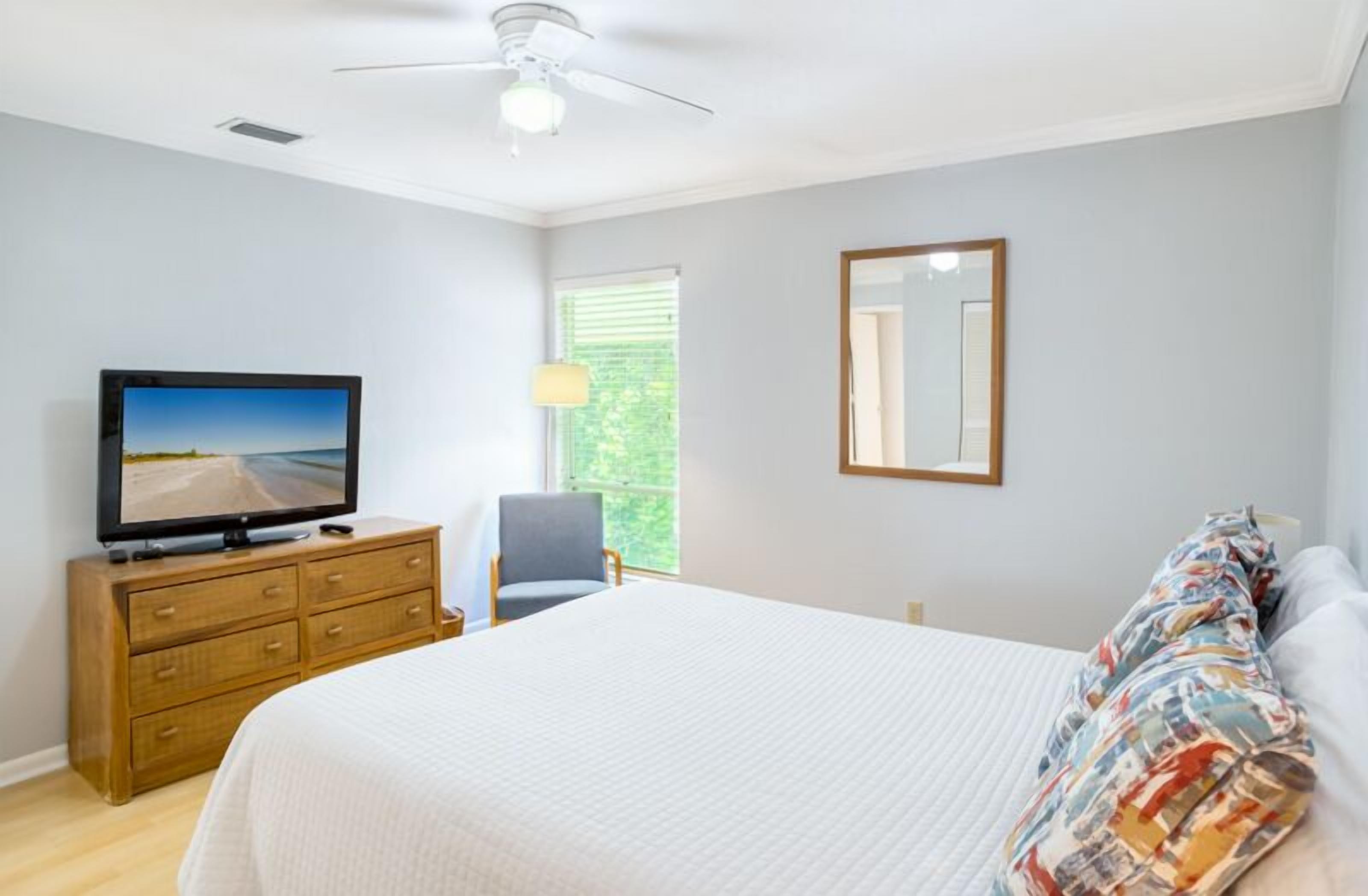 A bedroom with a bed, a dresser, a television, a ceiling fan, a window with blinds, and a mirror on the wall.