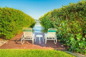 A wooden deck with two white lounge chairs and a small table, surrounded by lush green foliage.