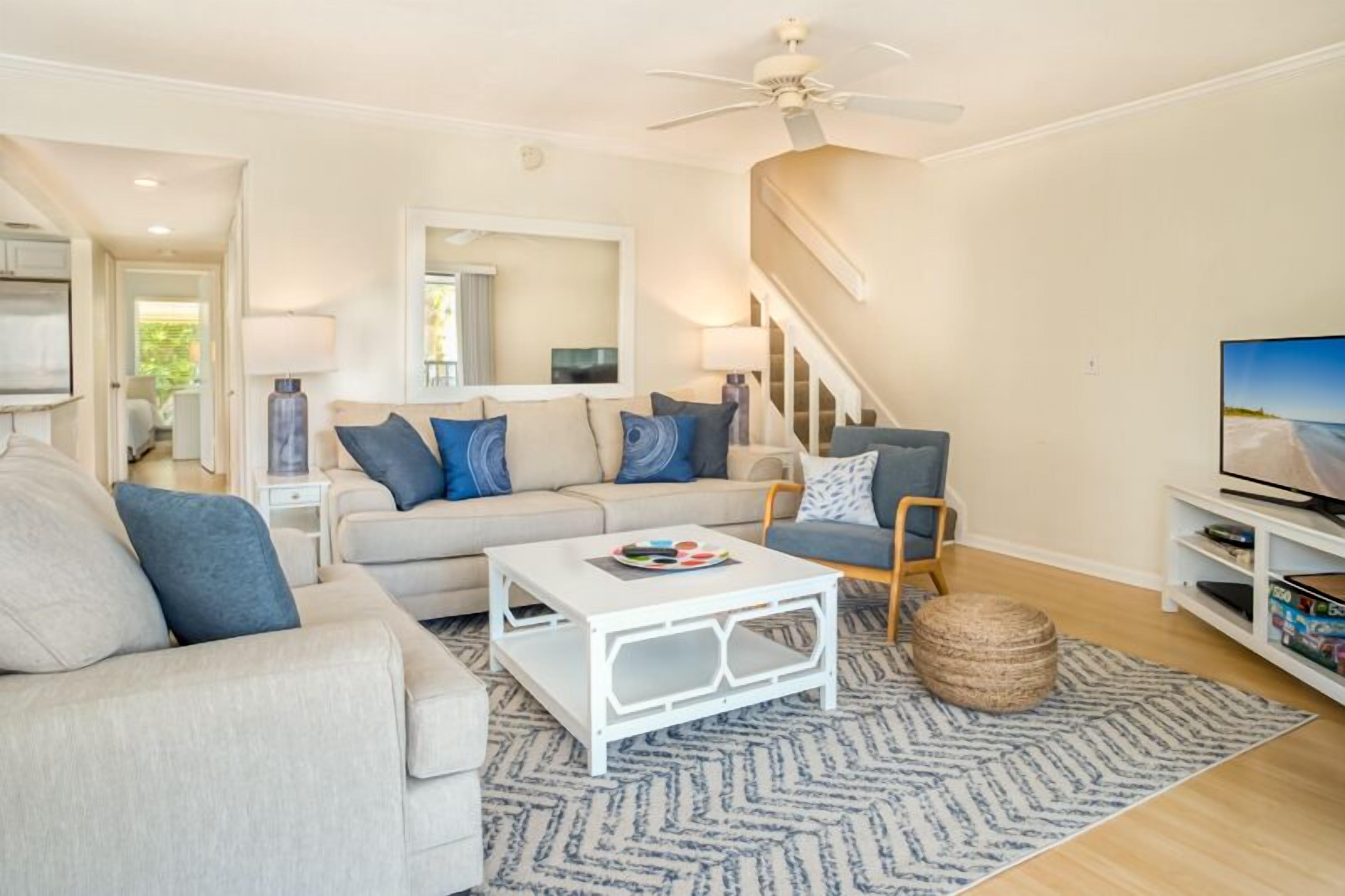 A living room with a sofa, coffee table, and TV. There is a staircase and a mirror on the wall.