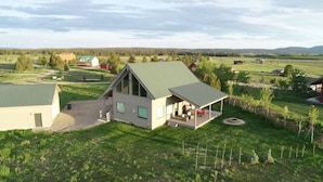 Una casa de dos pisos con techo verde, un porche con mobiliario exterior y un ventanal grande.
