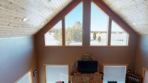Una habitación con chimenea de piedra, un televisor montado encima y amplias ventanas con vistas a un paisaje nevado.