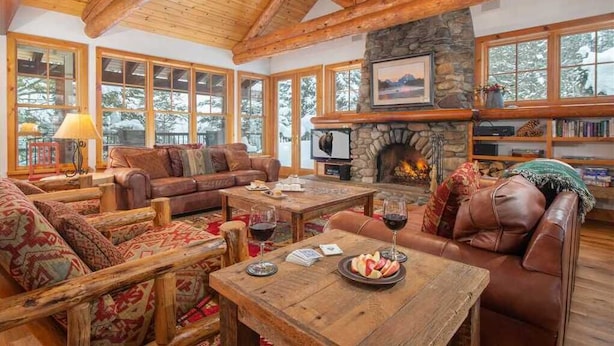 Un salon chaleureux avec des poutres en bois, une cheminée en pierre, des meubles en cuir et de grandes fenêtres.