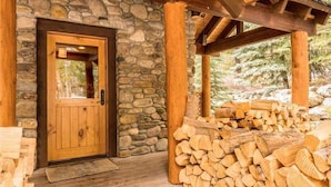 L’entrée d’un chalet rustique, avec un mur en pierre et une porte en bois, une véranda en bois dotée d’un toit, et une pile de bois de chauffage à l’extérieur.