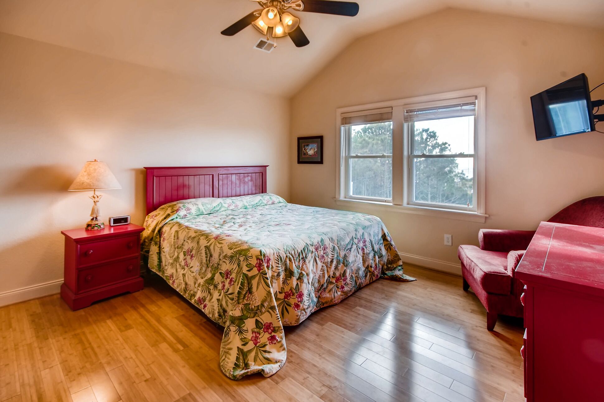 A bedroom with a wooden floor, a bed with a floral bedspread, a red nightstand with a lamp, a red chair, a ceiling fan, and a window with blinds.