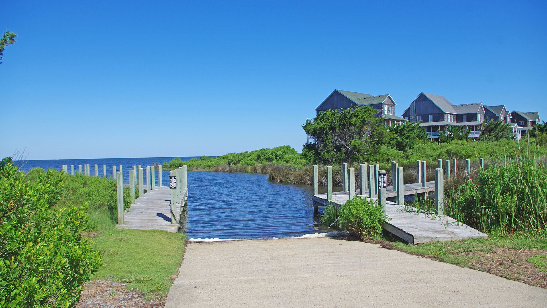 A waterfront property with a wooden dock leading to a pier, surrounded by lush greenery and a clear blue sky.