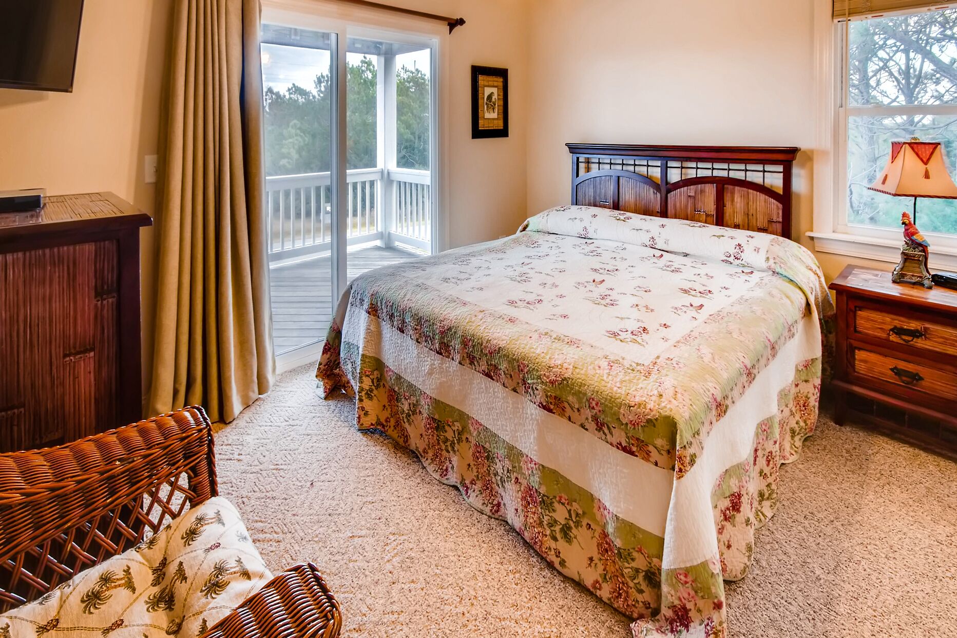 A bedroom with a bed, a wooden nightstand, a wicker chair, a wooden dresser, and a window with a view of trees.