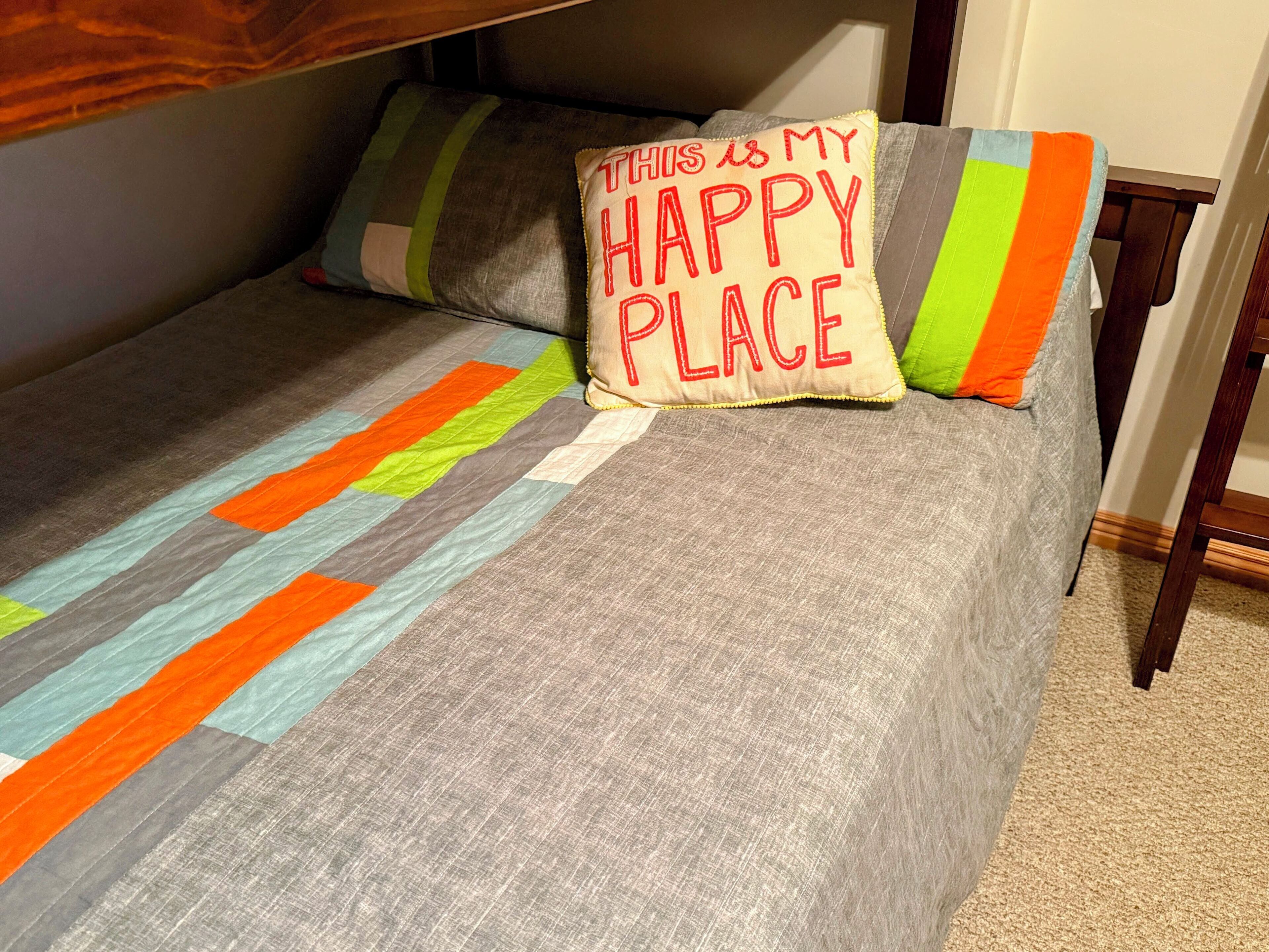 Ein Bett mit einer mehrfarbigen, gestreiften Decke und Kissen, eines davon mit dem Aufdruck "THIS IS MY HAPPY PLACE".