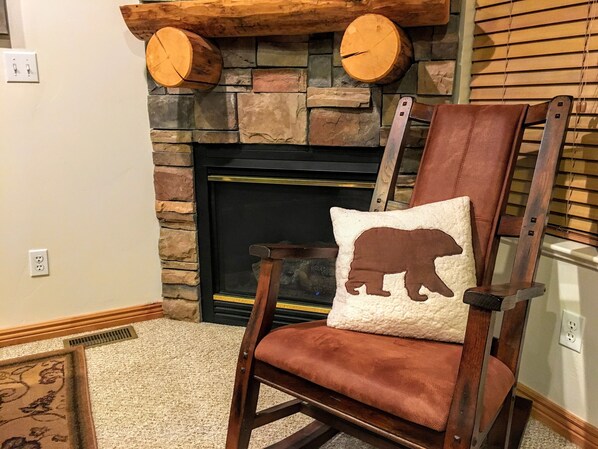 A wooden rocking chair with a bear-shaped pillow, positioned in front of a stone fireplace with two wooden logs. - SUMMER ALERT - Steps to Hot Tub - Family friendly (Eden)