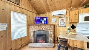 L’intérieur chaleureux d’un chalet avec une cheminée en pierre, des murs en bois, un téléviseur fixé au-dessus de la cheminée et un coin cuisine équipé d’un four à micro-ondes et d’un plan de travail.