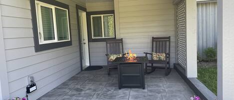 Ein überdachter Patio-Bereich mit Steinboden, zwei Holzstühle mit Kissen, einem kleinen Tisch mit Feuerstelle und Topfpflanzen.
